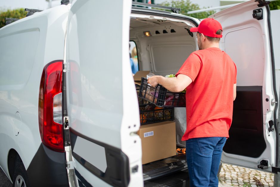 A courier in a red cap loads a delivery van with packages and a basket outdoors in Portugal.